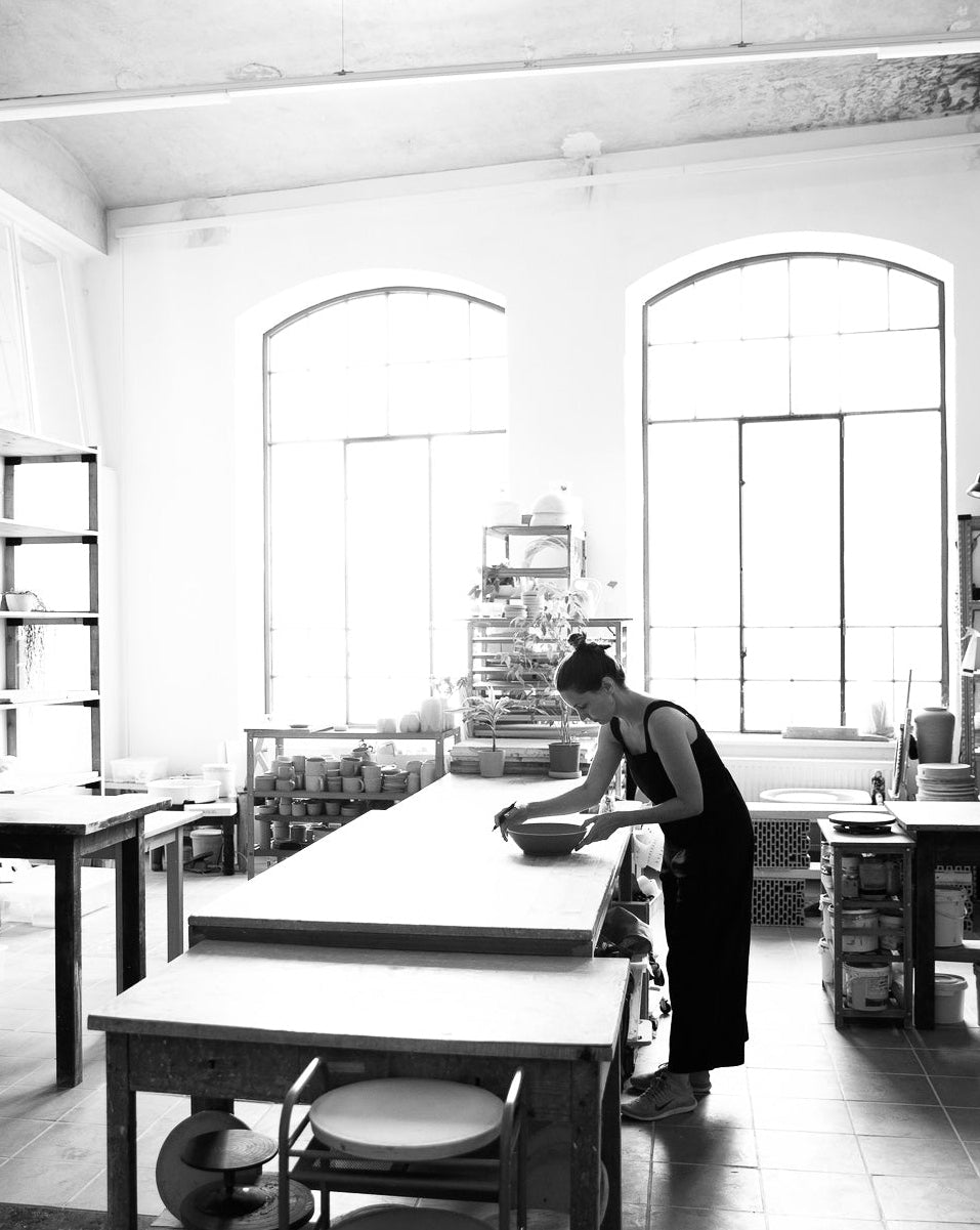 Die Keramikerin Serena Maria Zimmermann steht in ihrer Werkstatt an einem großen Arbeitstisch in der Mitte und bearbeitet eine lederharte Tonschüssel. An der Wand sind zwei große Fenster und an unterschiedlichen Stellen befinden sich Regale.
