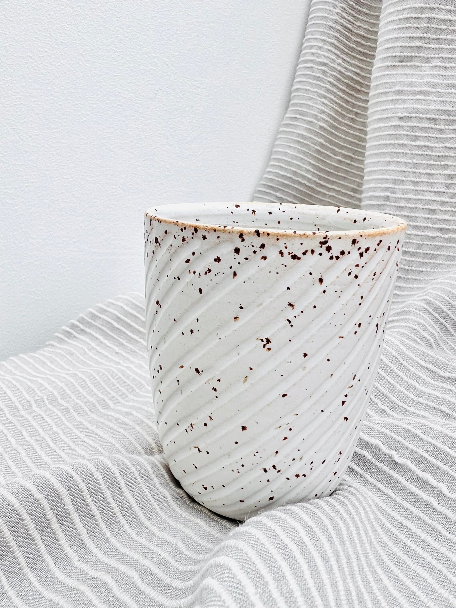 Ein Becher aus Steinzeug-Keramik mit geschwungenen Linien. Gesprenkelter Ton und mattweiße Glasur. Er steht auf einem grau-weiß gestreiften Tuch.