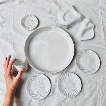 Sieben weiße Keramiken sind aufgestellt auf einem Tisch mit weißem Tischtuch. Eine Hand hält eine kleine Tasse. 