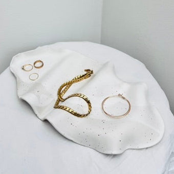 Ein weißes geschwungenes Brett steht auf einem kleinen Runden Tisch mit weißem Tischtuch. Auf dem brett liegen sechs Schmuckstücke. 