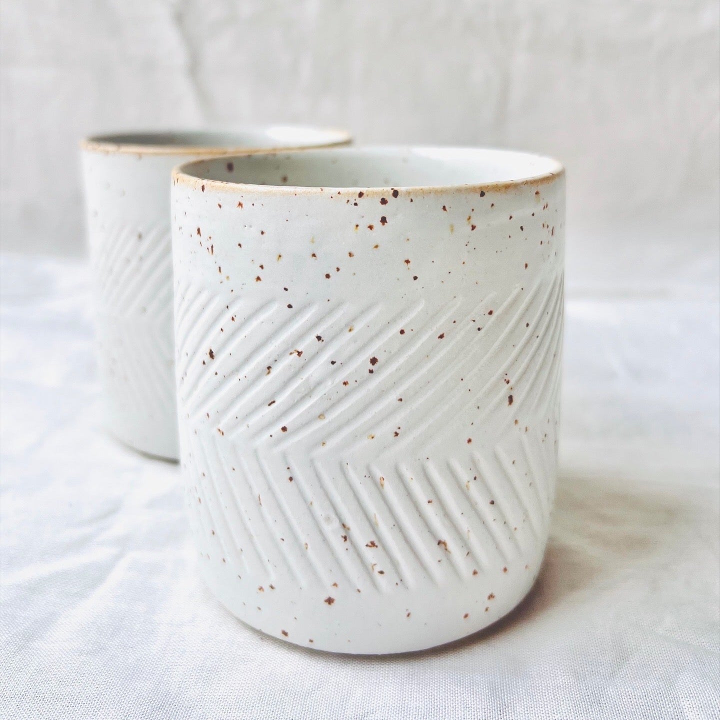 Zwei weiße Becher mit eingeschnitzten Zick Zack Muster stehen auf einem Tisch mit weißem Tischtuch und Hintergrund. 